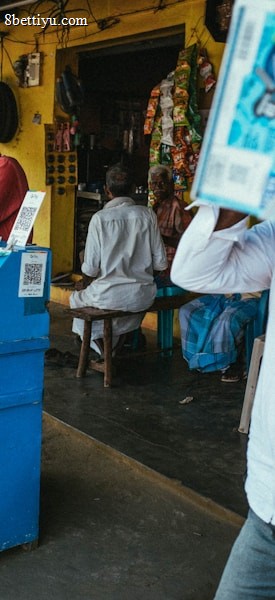 A street vendor stall displaying an array of lottery tickets clipped to a bright blue kiosk. A man in a red shirt leans on the counter, appearing thoughtful, while another man in a white shirt adjusts his hat and walks by. A variety of products hangs from strings above, and a colorful display fills the background with vibrant signs and packets. The scene suggests a busy, lively marketplace.